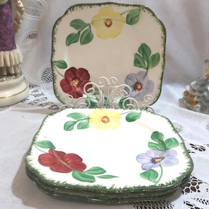 May include: A stack of square ceramic plates with a floral design. Each plate features a white background with a green rim and painted flowers in red, yellow, and purple, along with green leaves. A decorative plate holder is behind the stack.