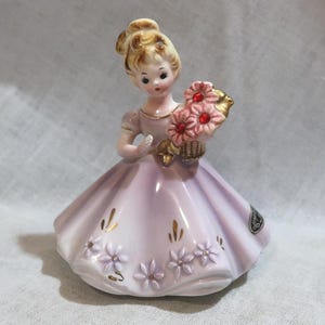 May include: A porcelain figurine of a young woman in a lavender dress, holding a basket of pink flowers. The figurine has blonde hair and is adorned with gold accents. The dress features floral and gold leaf details.
