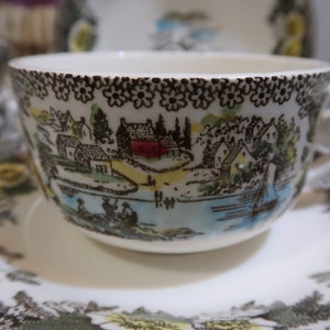 May include: A white teacup and saucer with a brown, green, and blue floral pattern. The pattern features a small village with houses, trees, and a river.