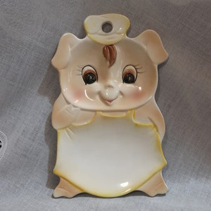 May include: A ceramic pig-shaped spoon rest with a yellow apron and brown hair. The pig has large eyes and a smiling mouth.