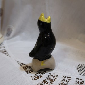 May include: A black ceramic bird with a yellow beak and crown, perched on a beige ceramic base. The bird is facing upwards with its beak open.