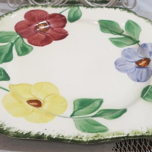 May include: Square ceramic plate with a cream-colored background, adorned with hand-painted flowers in red, yellow, and blue, along with green leaves. The plate has a scalloped edge with a green trim, creating a vintage aesthetic.
