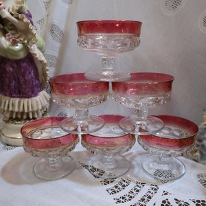 May include: Six vintage glass dessert dishes with a red and gold rim. The dishes are stacked in a pyramid shape on a white tablecloth.