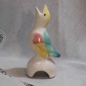 May include: A ceramic bird-shaped bottle pourer with a yellow beak, pink, blue, and yellow painted details, and a white base.