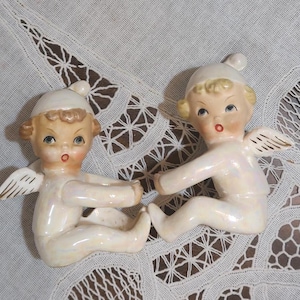 May include: Two ceramic angel figurines. Each angel is seated, wearing a white cap and outfit, with small gold-tipped wings. The angels have blonde hair, blue eyes, and rosy cheeks and lips. The figurines are on a white, patterned surface.