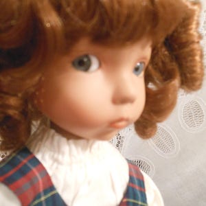 May include: A porcelain doll with auburn curly hair and blue eyes. The doll wears a white blouse under a plaid jumper in red, blue, and yellow. The doll's face has a delicate, painted expression.
