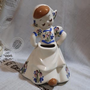 May include: A white ceramic figurine of a girl wearing a blue and white dress with floral accents. The girl has brown hair and blue eyes and is holding her skirt up to reveal a small vase.