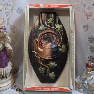 May include: A brown wooden wall plaque with a raised design of a copper teapot with green ivy vines. The plaque has the text "Wall Plaque For Your Home" printed on it.