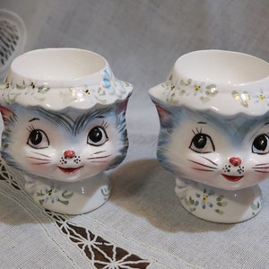 May include: Two blue and white ceramic egg cups shaped like cat heads with floral details. The cats have large eyes and a smiling expression.