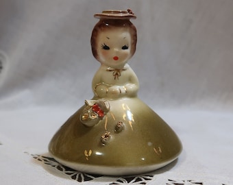 Może przedstawiać: Antyczna figurka porcelanowa przedstawiająca dziewczynkę w zielonej sukience i brązowym kapeluszu. Trzyma koszyk z kwiatami i ma słodki wyraz twarzy.