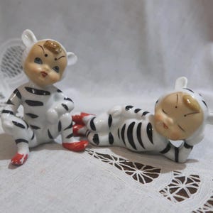 May include: Two vintage ceramic figurines of small children in white and black striped outfits. One is seated, the other is lying down. Both have red shoes and are wearing hats with white ears.