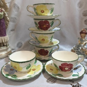 May include: A set of vintage teacups and saucers. The cups and saucers are white with hand-painted floral designs in yellow, red, and blue. Each piece has a green trim. The cups are stacked, and two sets are arranged in front.