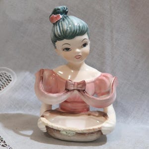 May include: A vintage ceramic figurine of a woman with a bun and a pink dress, holding a basket. The figurine has a delicate design with a bow detail on the dress. The woman's hair is a teal color.