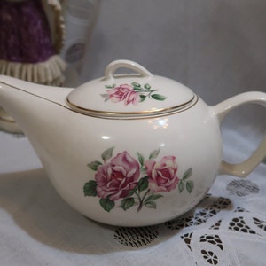 May include: White ceramic teapot with a floral design featuring pink roses and green leaves. The teapot has a gold trim around the lid and spout.