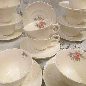 May include: A collection of cream-colored porcelain teacups and saucers featuring scalloped edges, embossed textured patterns, and delicate floral motifs of pink roses with green leaves painted on the interior and exterior surfaces.