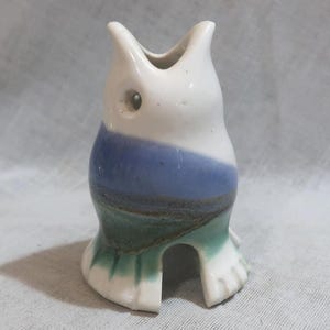 May include: A ceramic owl-shaped vase with a white head and a body featuring blue and green bands. The vase has a small opening at the top and a cutout base, showcasing a unique, handcrafted design. The owl has a small eye.