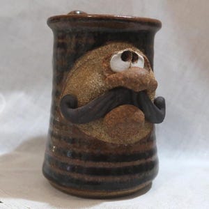 May include: A handmade ceramic mug with a whimsical face design. The mug features a brown and tan color scheme with a sculpted face, including a mustache and eyes. The mug has a cylindrical shape with a handle.