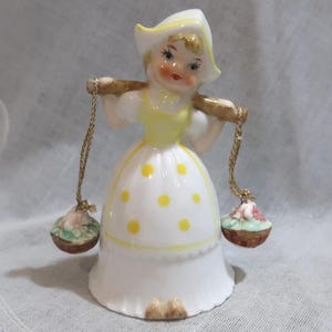 May include: A ceramic figurine of a young girl carrying two baskets. She wears a white dress with yellow polka dots and a yellow bodice. The girl has a white hat and is holding a wooden pole with gold chains attached to the baskets.