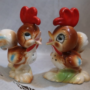 May include: A pair of ceramic rooster salt and pepper shakers. The rooster on the left is brown with blue accents and has a red comb. The rooster on the right is brown with green accents and has a red comb.