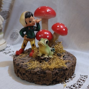 May include: A whimsical figurine set featuring a green frog, a small elf with a red hat and green clothing, and three red mushrooms with white polka dots. The figurines are arranged on a cork base with brown moss.