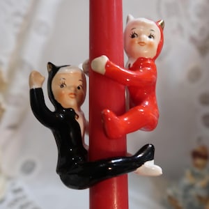 May include: Two porcelain figurines of devils climbing a red candle. One devil is wearing a red suit and the other is wearing a black suit.