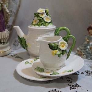 May include: A white ceramic teapot and creamer set with floral accents. The teapot has a lid with a floral design and a handle. The creamer has a handle and a floral design on the side. The set is on a white lace tablecloth.