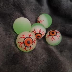 May include: A collection of five novelty eyeball-shaped balls. Four balls have a white base with a red vein pattern and a brown and orange iris. One ball is solid green. The balls are on a gray, textured surface.