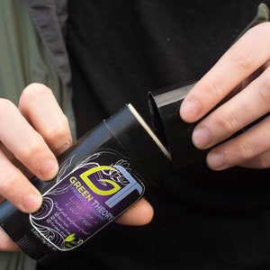 May include: A black and purple deodorant stick with the text "GREEN THEORY" and "Lavender Green" printed on the label. The deodorant is being held in a person's hand and the cap is being removed.