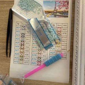 May include: Diamond painting kit with a printed guide featuring a color chart and a picture of the Eiffel Tower. Includes a tray of light blue rhinestones, a stylus, and tweezers. The kit is designed for crafting.