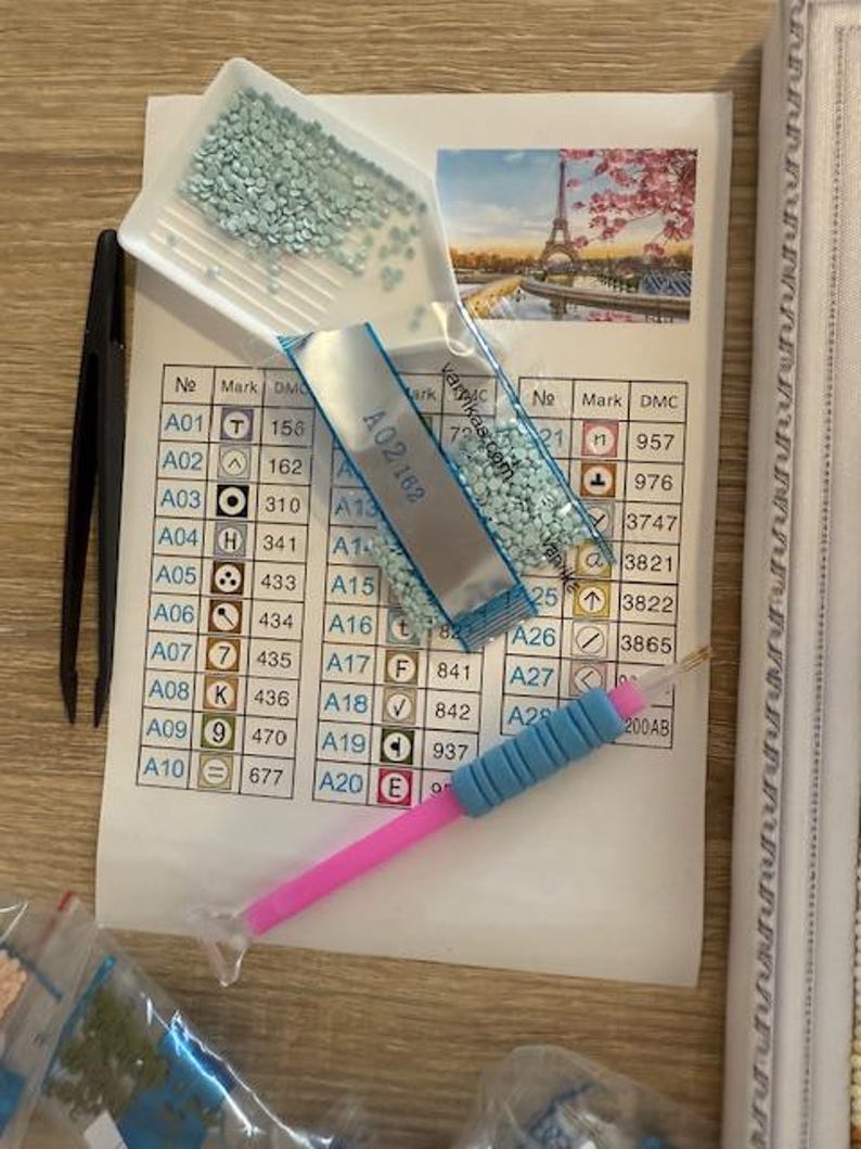 May include: Diamond painting kit with a printed guide featuring a color chart and a picture of the Eiffel Tower. Includes a tray of light blue rhinestones, a stylus, and tweezers. The kit is designed for crafting.