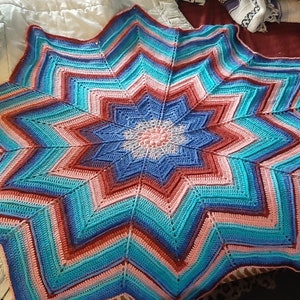 May include: A large crocheted star-shaped blanket in shades of pink, blue, and red. The blanket is made with a variety of stitches and has a textured look.