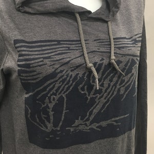 May include: A gray hooded sweatshirt with a dark blue graphic design on the front. The graphic appears to be a stylized landscape or mountain scene. The sweatshirt has a drawstring and a hood.
