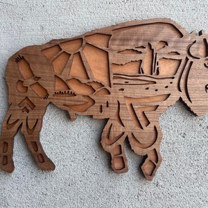 May include: A wooden cutout of a bison with intricate details, including a mountain range, trees, and a bird. The bison is brown and has a natural wood finish.