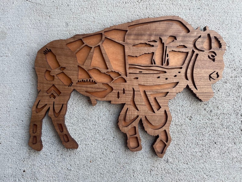 May include: A wooden cutout of a bison with intricate details, including a mountain range, trees, and a bird. The bison is brown and has a natural wood finish.