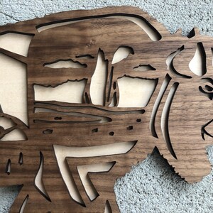 May include: A wooden wall art piece depicting a bison with a detailed silhouette of a landscape in the background. The wood is a dark brown color and the piece is cut out in a geometric pattern.