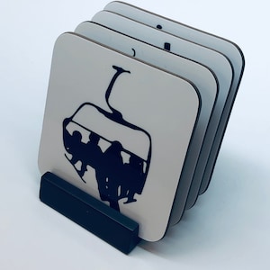 May include: Set of four white coasters with a black silhouette of a ski lift with people in it. The coasters are stacked and sitting on a black stand.