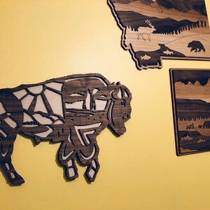 May include: A wooden wall art piece depicting a bison with a detailed landscape design. The bison is brown and the landscape is a light brown color.