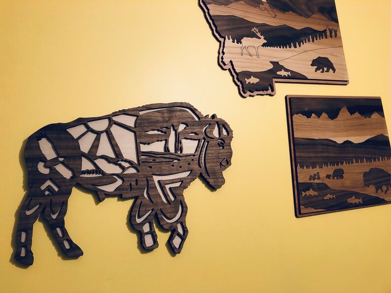 May include: A wooden wall art piece depicting a bison with a detailed landscape design. The bison is brown and the landscape is a light brown color.