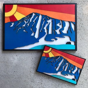 May include: Two wood wall art pieces depicting a mountain range with a sunset in the background. The larger piece is framed in black and the smaller piece is unframed. Both pieces are painted in shades of blue, red, orange, and yellow.