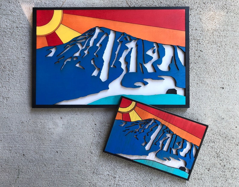 May include: Two wood wall art pieces depicting a mountain range with a sunset in the background. The larger piece is framed in black and the smaller piece is unframed. Both pieces are painted in shades of blue, red, orange, and yellow.