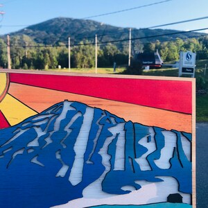 May include: A wooden sign with a stylized image of a mountain with a blue, white, and orange color scheme. The sun is depicted in the upper left corner with yellow and orange rays. The sign is in front of a mountain range.