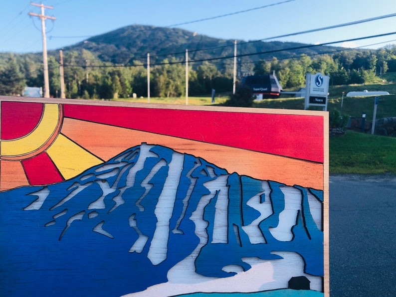 May include: A wooden sign with a stylized image of a mountain with a blue, white, and orange color scheme. The sun is depicted in the upper left corner with yellow and orange rays. The sign is in front of a mountain range.