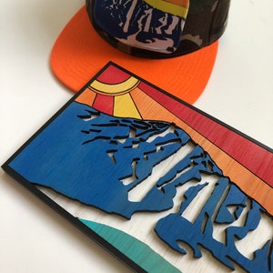 May include: A wooden wall art piece featuring a stylized mountain range with a sunburst design. The mountain range is blue, with a teal and white background. The sunburst is yellow, orange, and red. The piece is framed in black.