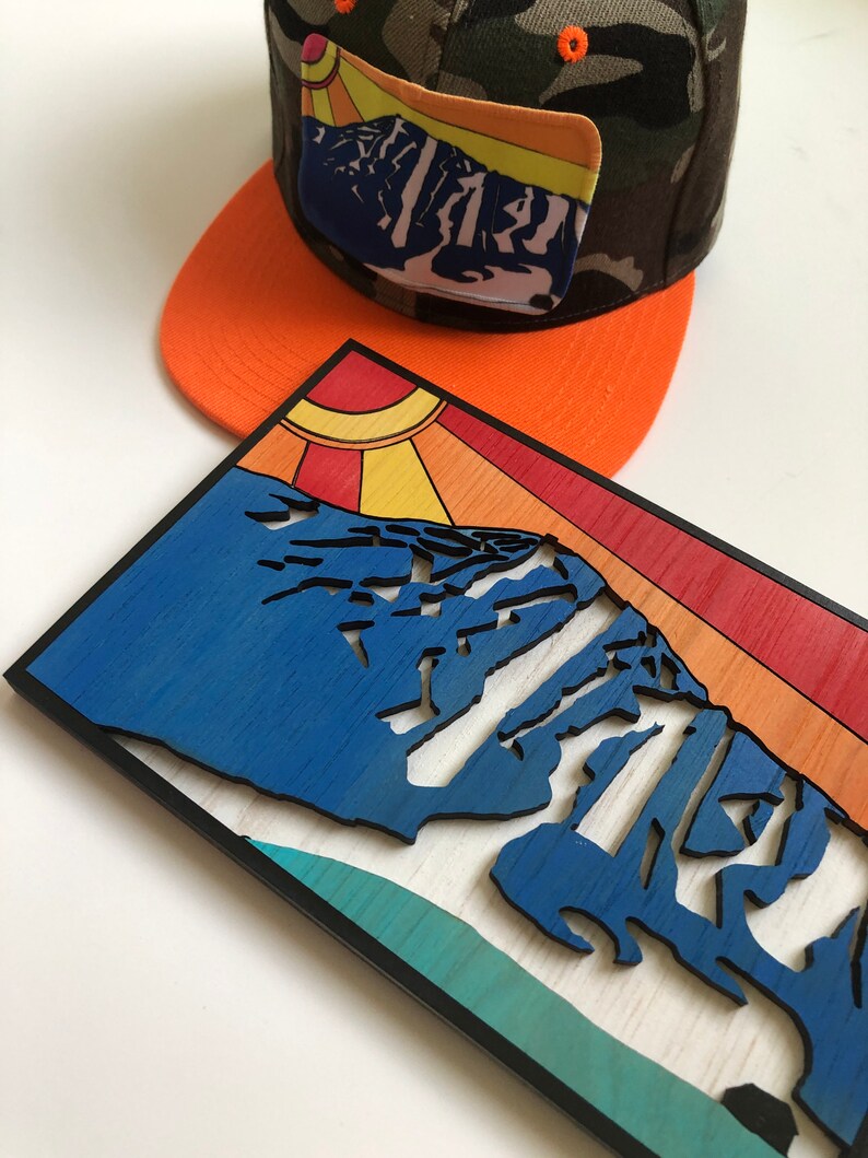 May include: A wooden wall art piece featuring a stylized mountain range with a sunburst design. The mountain range is blue, with a teal and white background. The sunburst is yellow, orange, and red. The piece is framed in black.