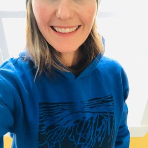 May include: A woman wearing a blue hoodie with a black graphic design and a gray beanie. The hoodie has a hood and a front pocket. The graphic appears to be an abstract landscape. The woman is smiling.