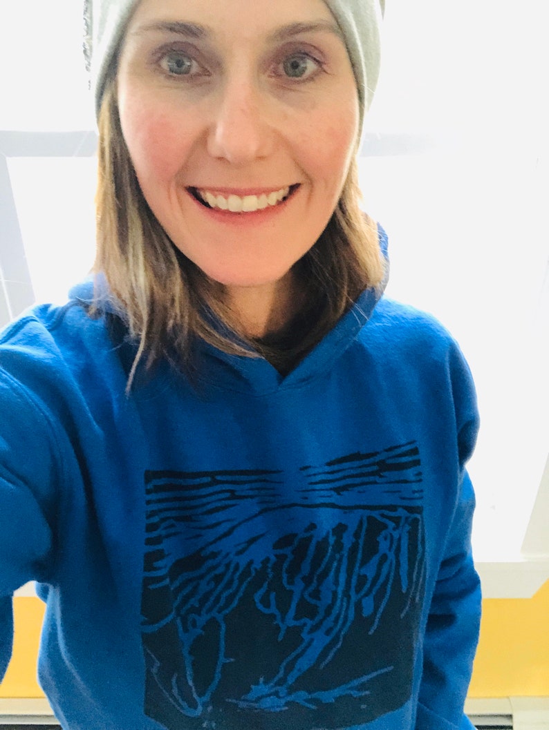 May include: A woman wearing a blue hoodie with a black graphic design and a gray beanie. The hoodie has a hood and a front pocket. The graphic appears to be an abstract landscape. The woman is smiling.