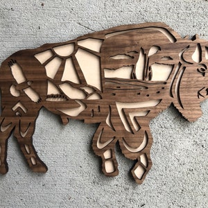 May include: A wooden wall art sculpture of a bison with a detailed design of a landscape scene inside the silhouette. The bison is made of dark wood and the landscape is cut out to reveal a lighter wood underneath.