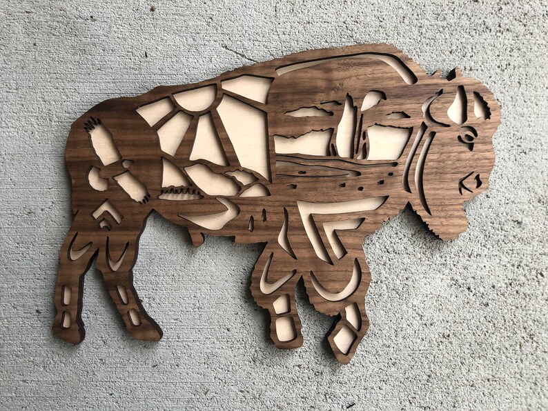 May include: A wooden wall art sculpture of a bison with a detailed design of a landscape scene inside the silhouette. The bison is made of dark wood and the landscape is cut out to reveal a lighter wood underneath.