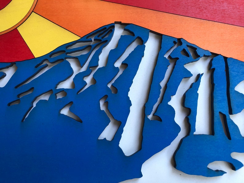 May include: A blue wooden mountain with white cutouts, resembling a ski slope. The mountain is set against a background of orange, red, and yellow.
