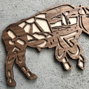 May include: A wooden wall art sculpture of a bison with intricate geometric patterns and a stylized landscape design. The bison is brown and the background is a light wood color.